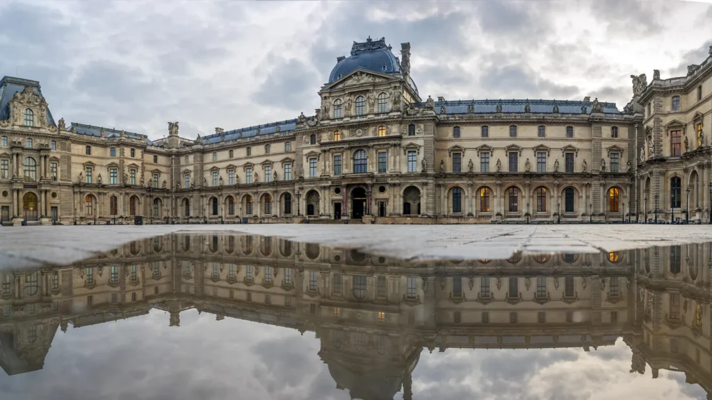 The Louvre palace facade and cloudy sky are reflected in water in the courtyard.