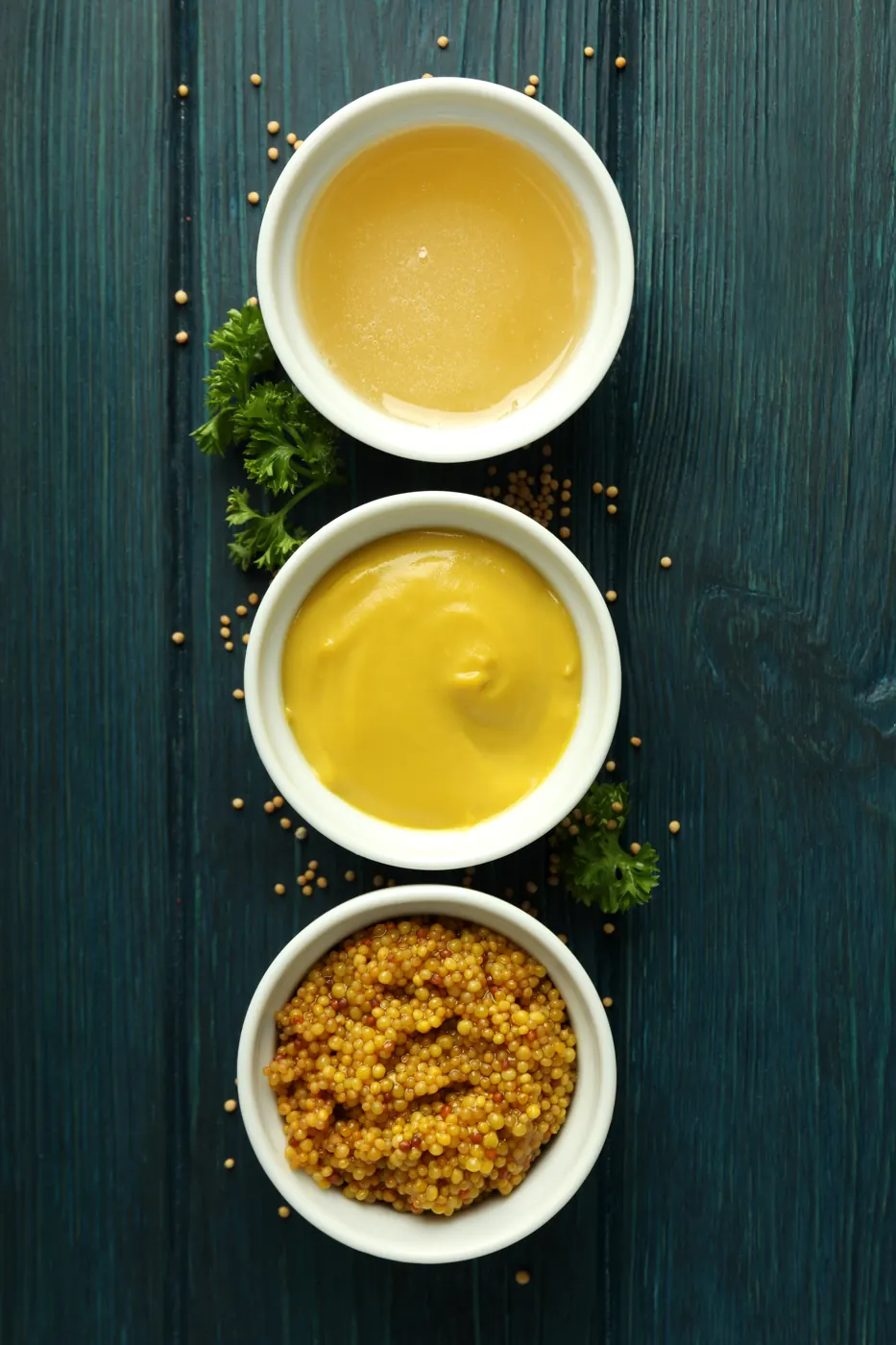Three white bowls holding smooth and grainy prepared mustards on a blue wooden surface.
