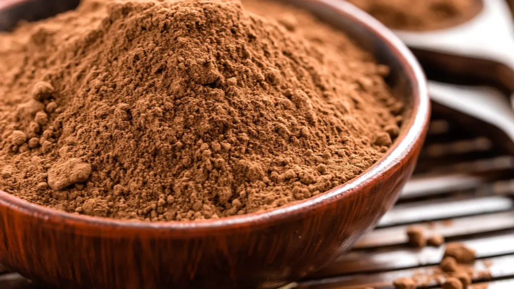 A wooden bowl heaped with cocoa powder beside chocolate pieces and a spoon.