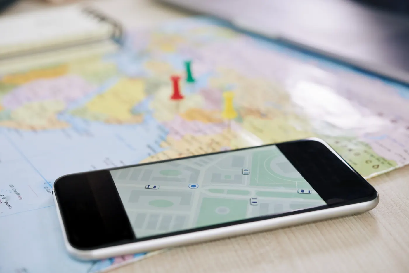 A smartphone displaying a street map rests on a paper map beside colored pins.