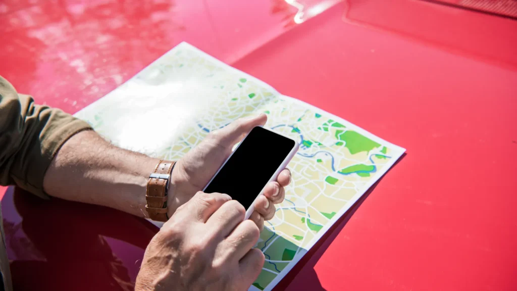 Hands hold a smartphone above a paper map on the red hood of a car.