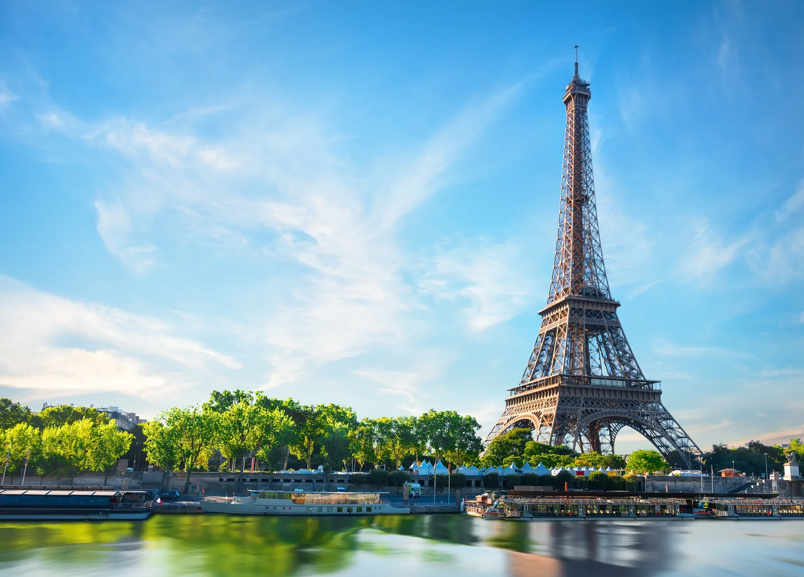 The Eiffel Tower rises beside the Seine and a bridge on a bright day in Paris.