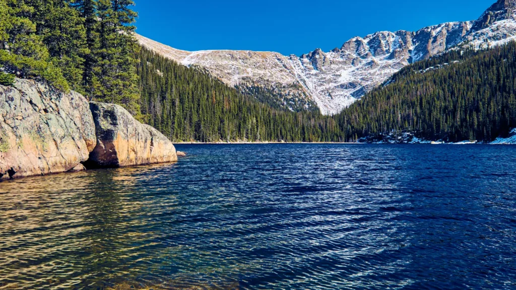 Blue lake water lies below evergreen slopes and a snow-streaked mountain ridge at Lake Verna, Colorado.