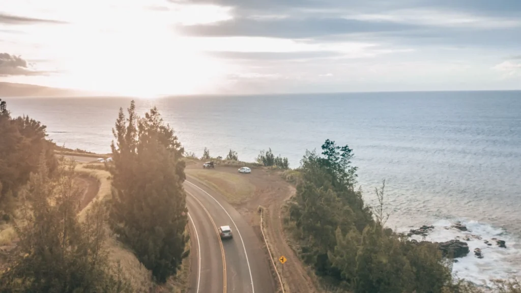 A car following a curving coastal road beside the ocean at sunset.