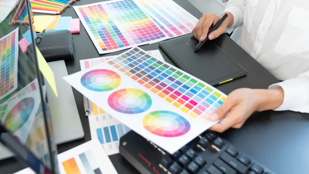 A designer using a pen tablet beside printed color charts and a computer.
