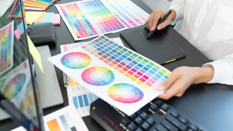 A designer using a pen tablet beside printed color charts and a computer.