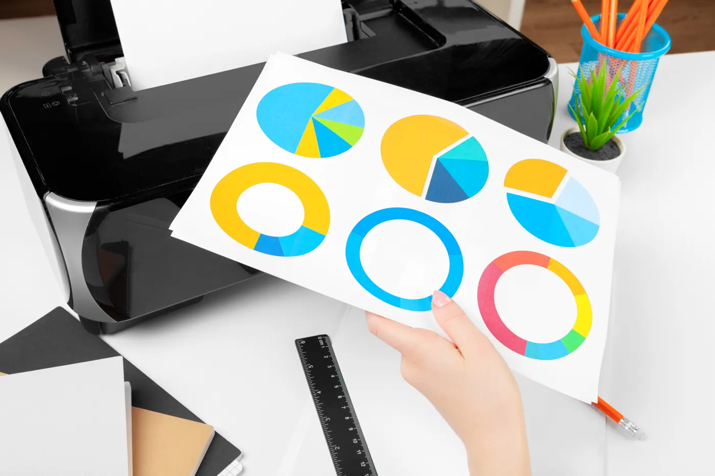 A hand holding a printed page of colored charts in front of a desktop printer.