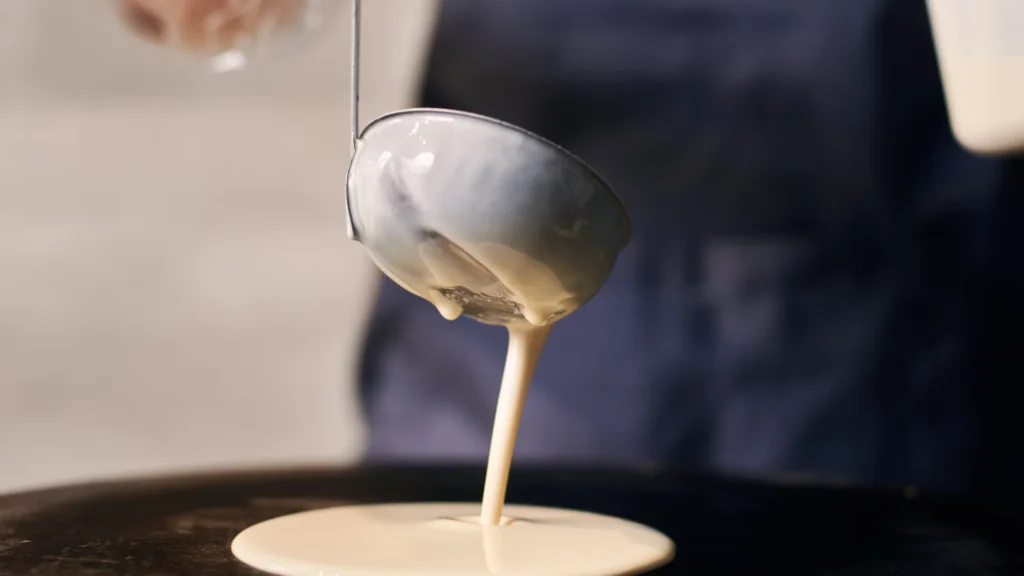 A ladle pours pancake batter onto a round cooking surface.