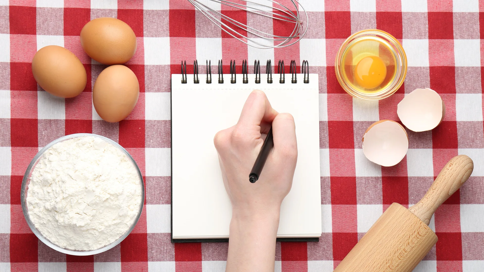 A hand holding a pen over a blank notebook beside eggs, flour, and baking tools.