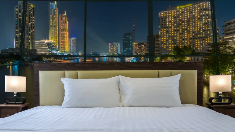 A neatly made hotel bed facing large windows overlooking illuminated riverside buildings at night.