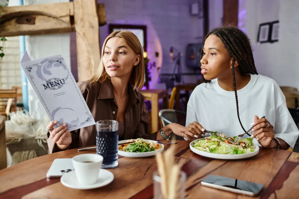 Two diners at a restaurant table looking toward a menu, with salads and drinks in front of them.
