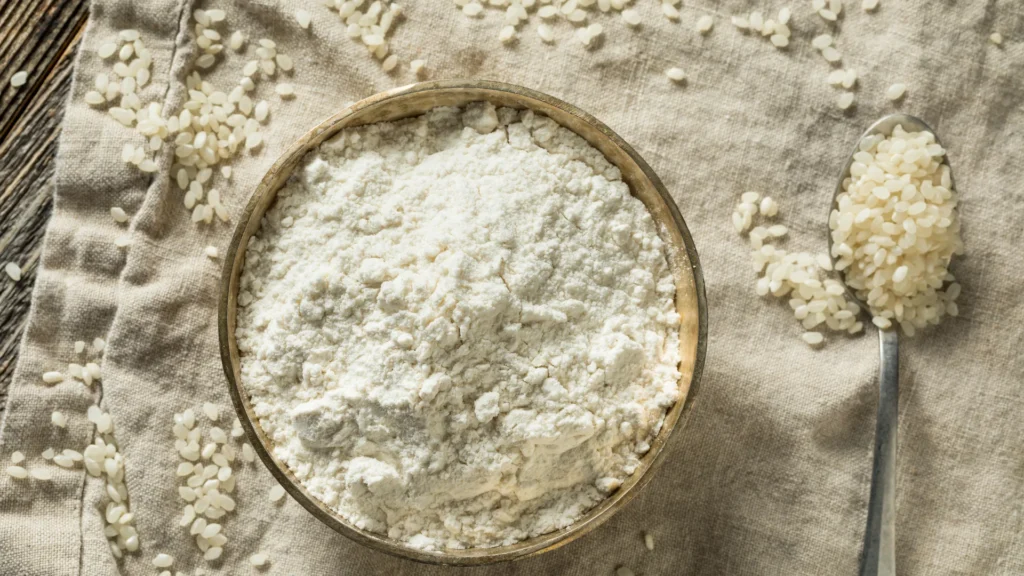 A bowl of rice flour surrounded by rice grains and a metal scoop.