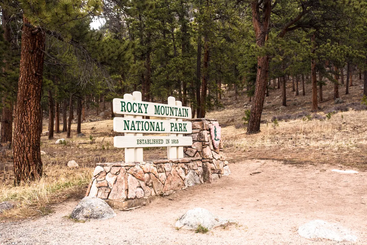 A sign reading Rocky Mountain National Park stands on a stone base beside a wooded path.