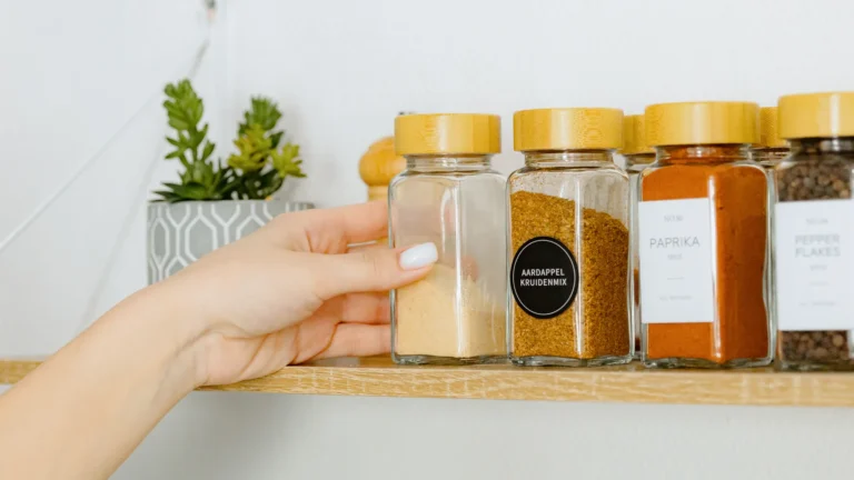 A hand reaching for a glass spice jar beside labeled jars on a kitchen shelf.