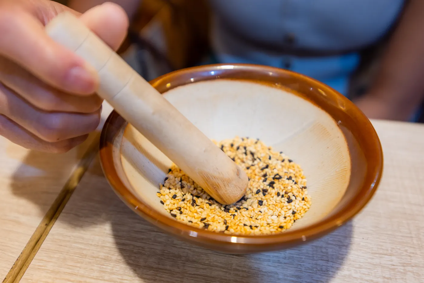A hand uses a pestle to grind a sesame seed mixture in a grooved ceramic mortar.