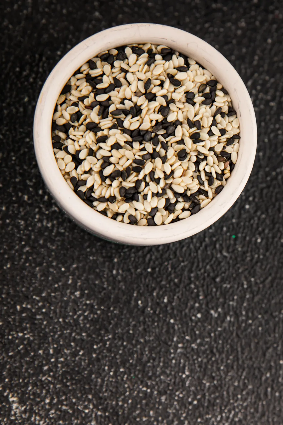 A small white bowl containing black and white sesame seeds on a dark surface.