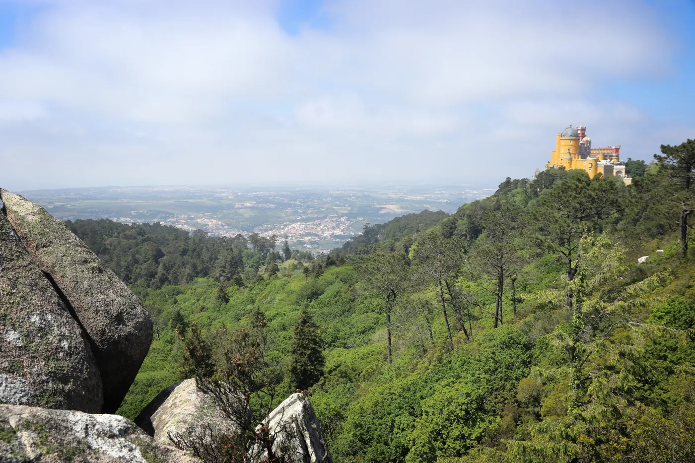 Pena Palace on a distant wooded hill with rocks and green slopes in the foreground