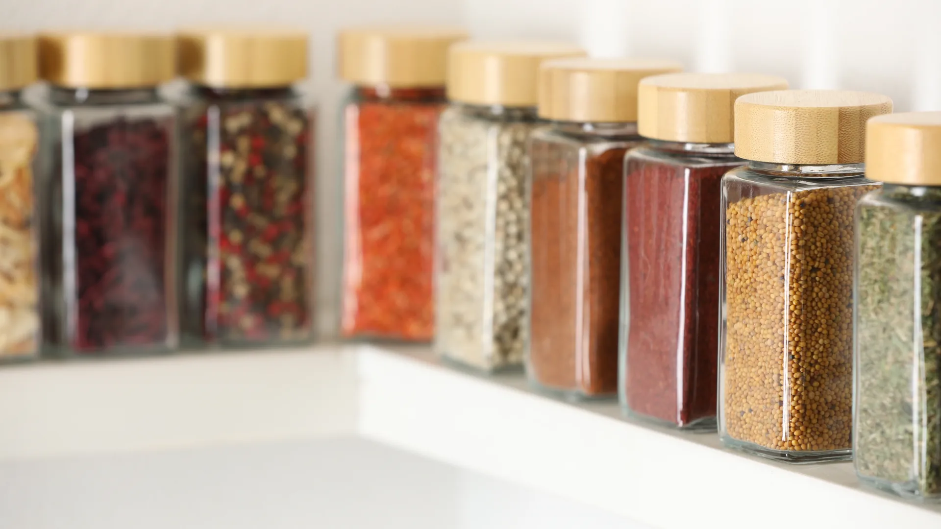 Closed glass spice jars with wooden lids arranged on a white shelf.