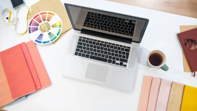 An open laptop with a color wheel, colored paper, notebooks, and a tape measure on a desk.