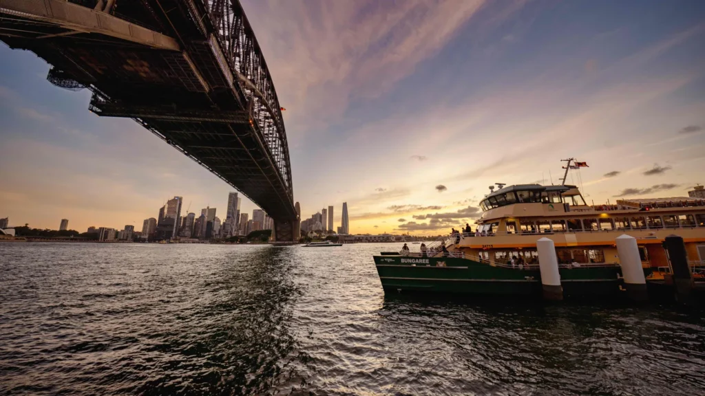 Green and yellow ferry beside the Sydney Harbour Bridge at sunset, with city buildings across the water