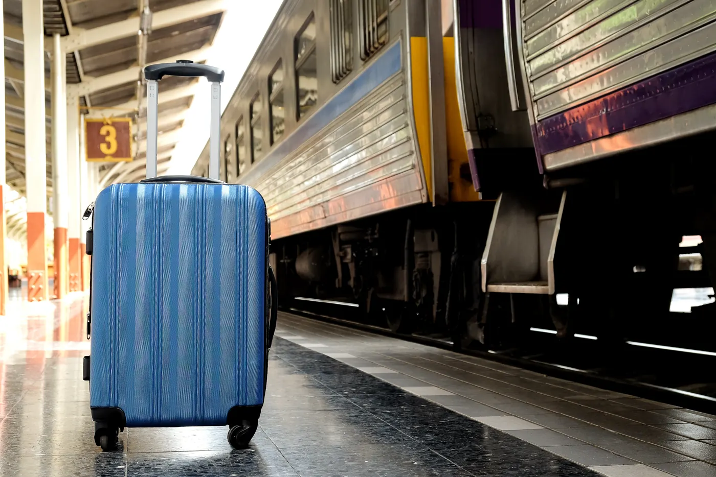 A blue rolling suitcase on a platform beside a passenger train.