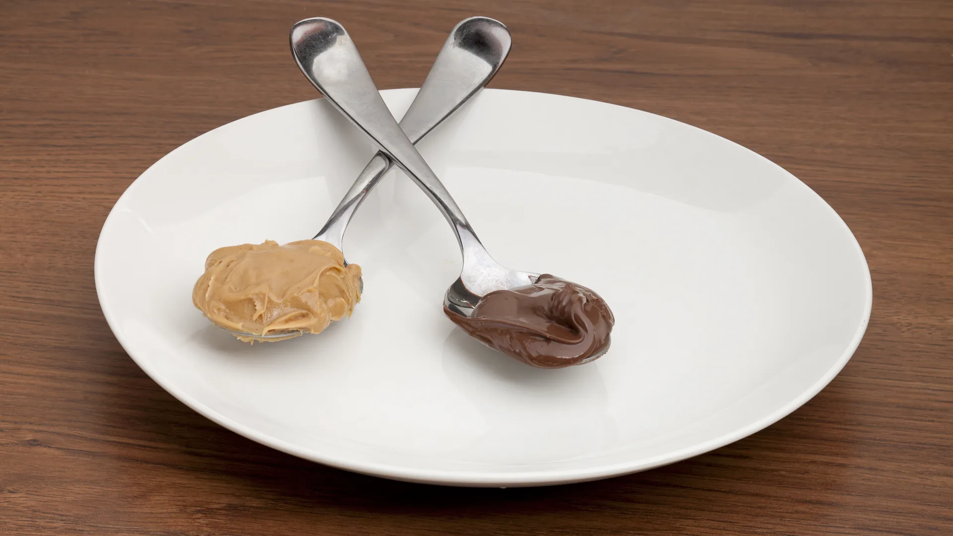 Two spoons holding peanut butter and chocolate spread on a white plate.