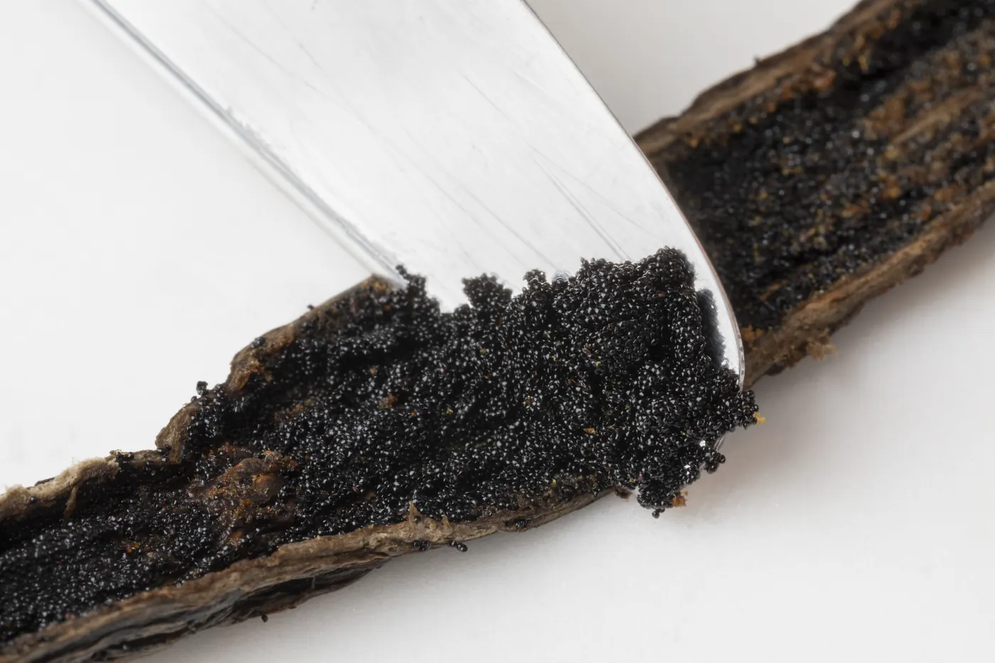 A knife lifts black vanilla seeds from a split vanilla bean on a white surface.
