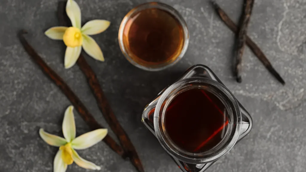 Dark vanilla extract in glass containers sits with whole vanilla beans and pale yellow flowers on a gray surface.