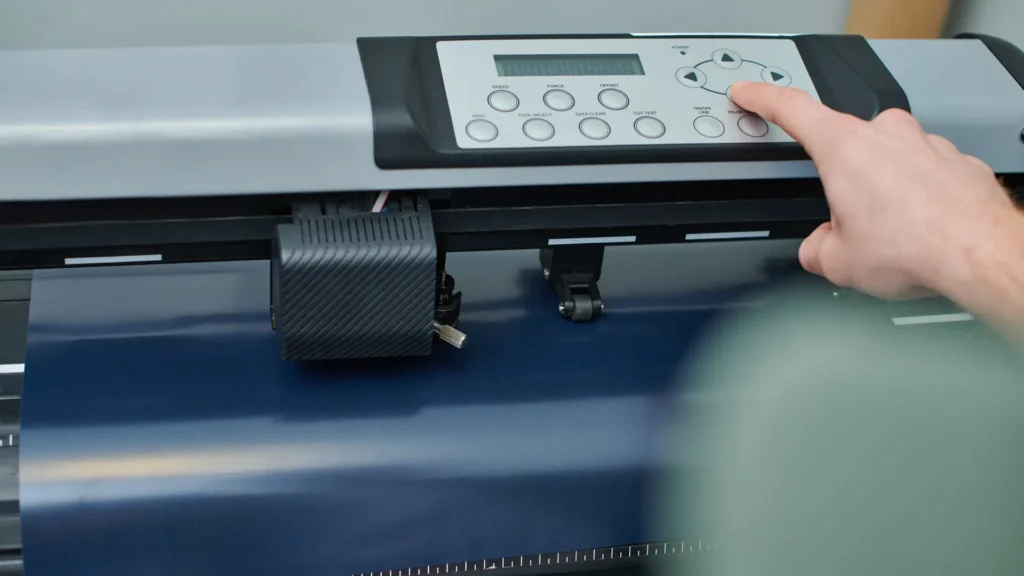 A hand at the controls of a cutting plotter with blue material loaded