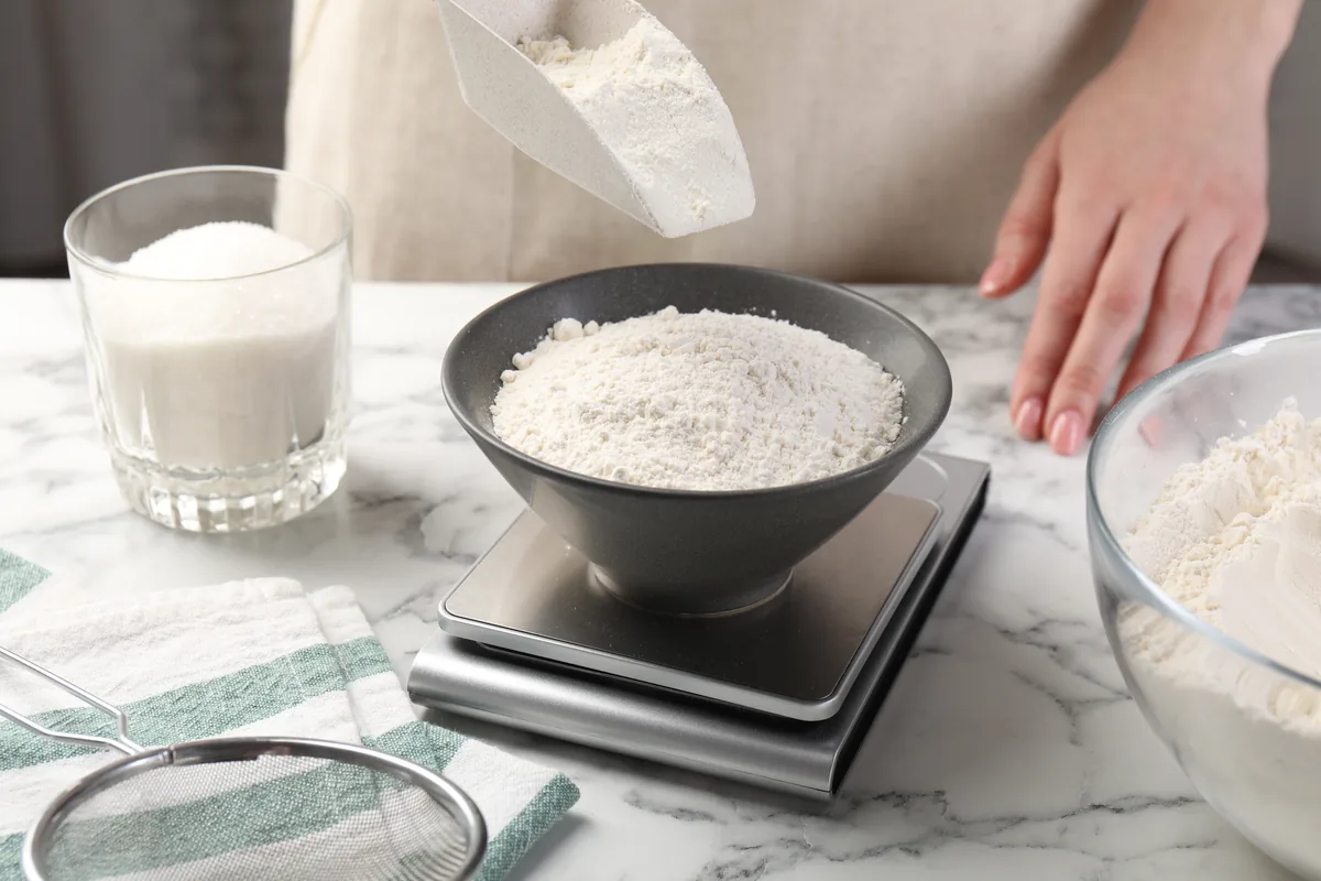 Flour being added to a bowl on a kitchen scale, with other baking ingredients nearby.