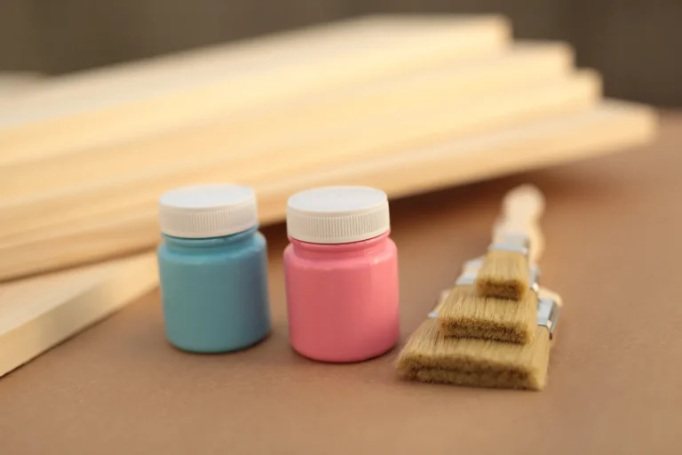 Blue and pink paint jars with wooden-handled brushes and unfinished wooden pieces on a work surface.