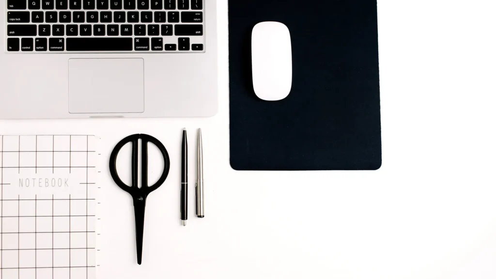Overhead view of a laptop, gridded notebook, black scissors, pens, and mouse on a white desk
