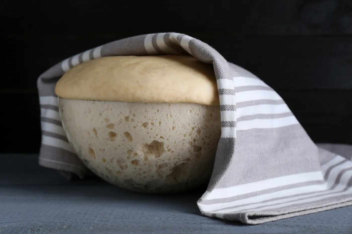 Expanded bread dough rising above a clear bowl partly covered with a striped towel